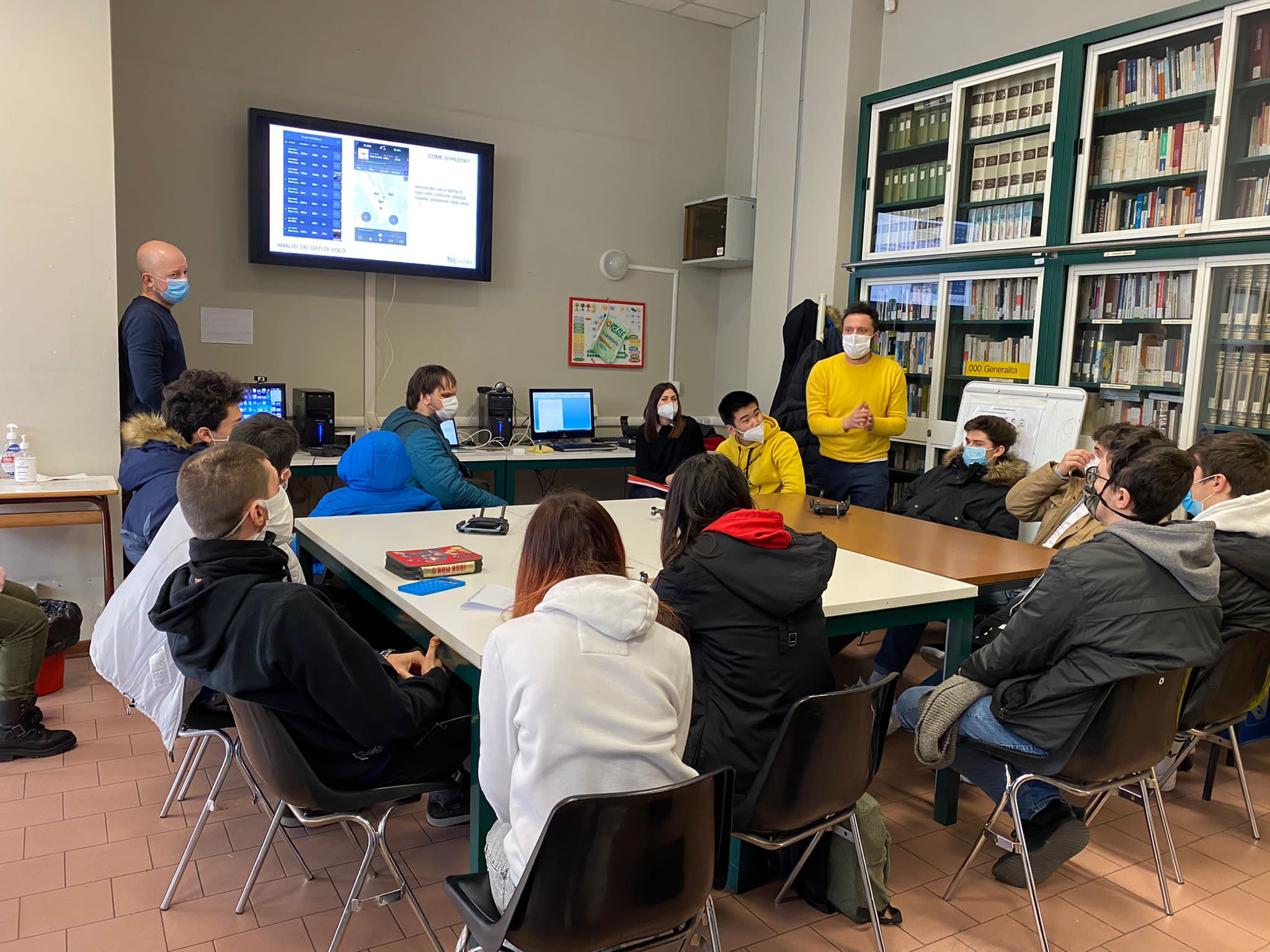 Students sitting at a table listening to a teacher speaking.