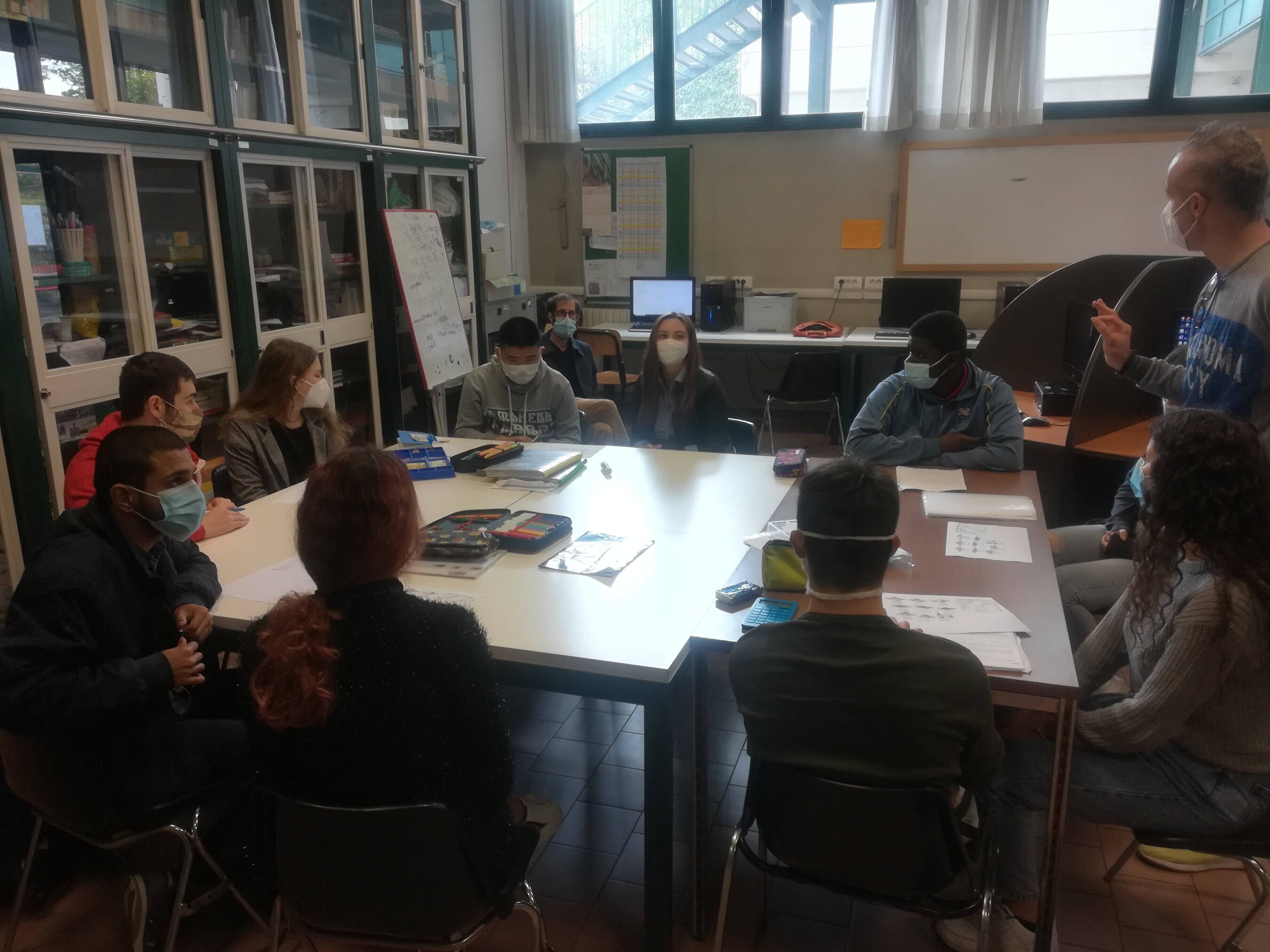 Students sitting at a table listening to a teacher speaking.