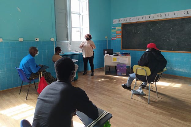 A woman in a mask stands in a classroom in front of four men seated at desks