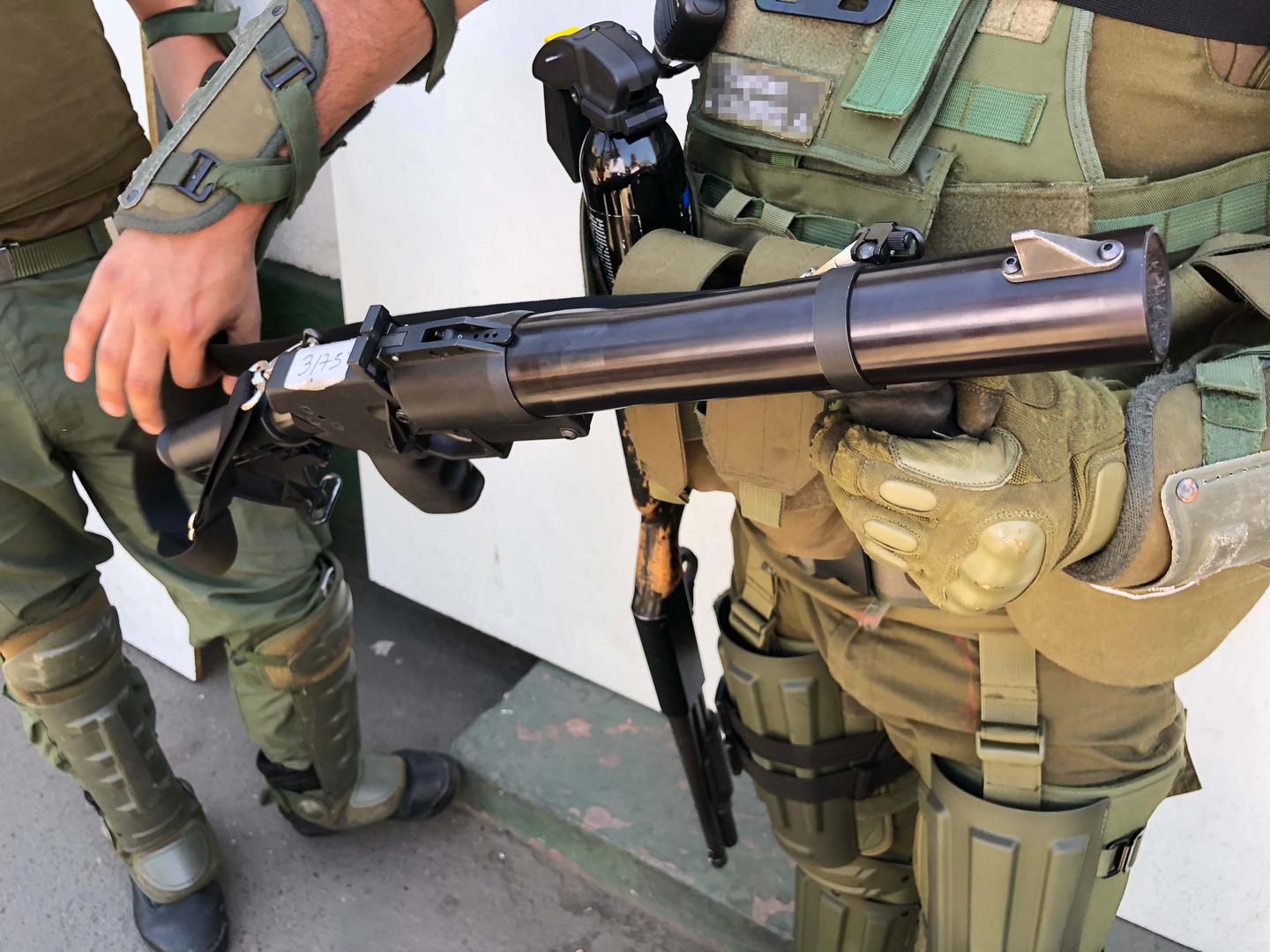 A police captain holds a teargas gun. A pellet shotgun hangs on his right side. 