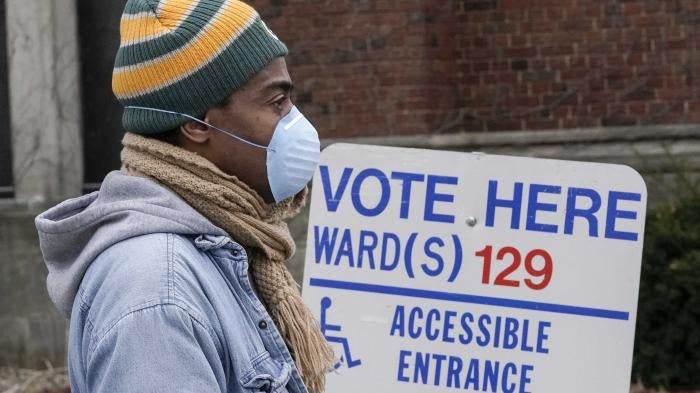 A man in a beanie and face mask stands in front of a sign that says "Vote Here"