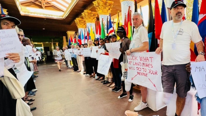 People protest in a corridor with two men with amputated legs hold a sign that says "Look what AP landmines will do to your people."