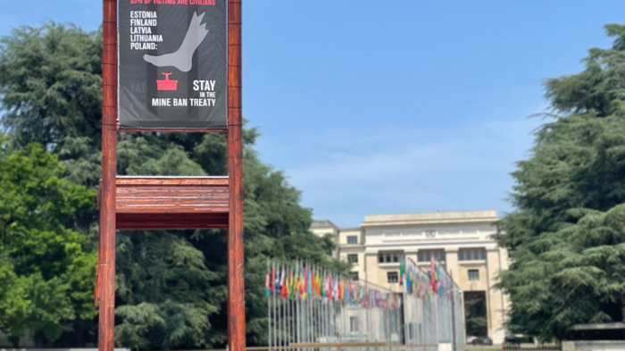 A banner hangs on the Broken Chair sculpture stating "Landmines don't choose their targets. 85% of victims are civilians. Estonia, Finland, Latvia, Lithuania, and Poland stay in the Mine Ban Treaty."