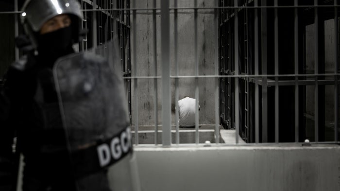 A person sits inside a cell while a guard with a riot shield stands outside