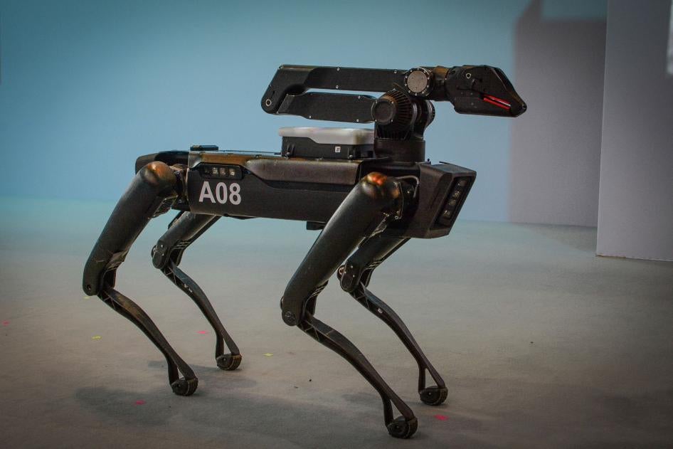 A black robot with four legs resembling a dog stands on a stage in front of a shite background.
