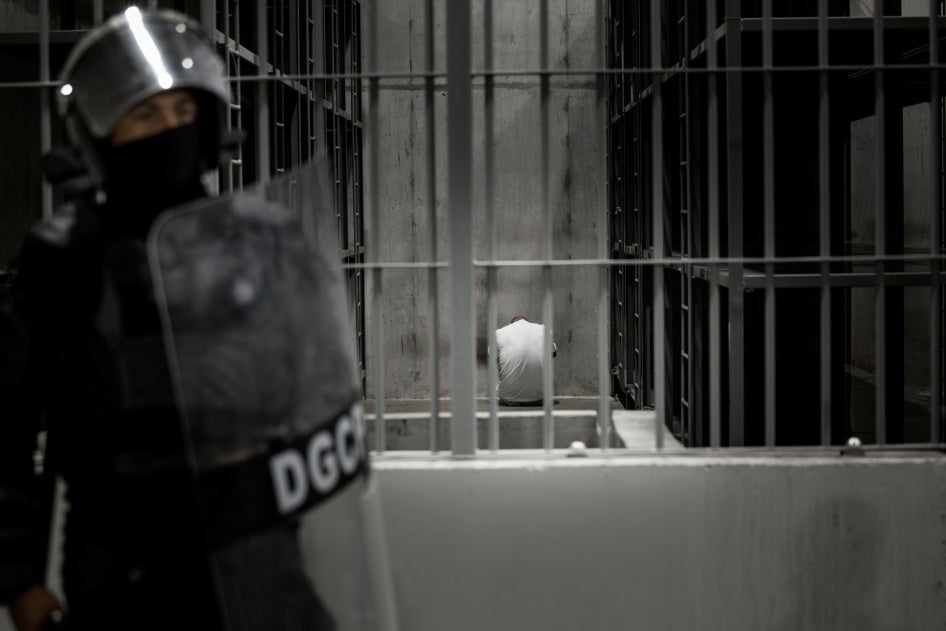 A person sits inside a cell while a guard with a riot shield stands outside