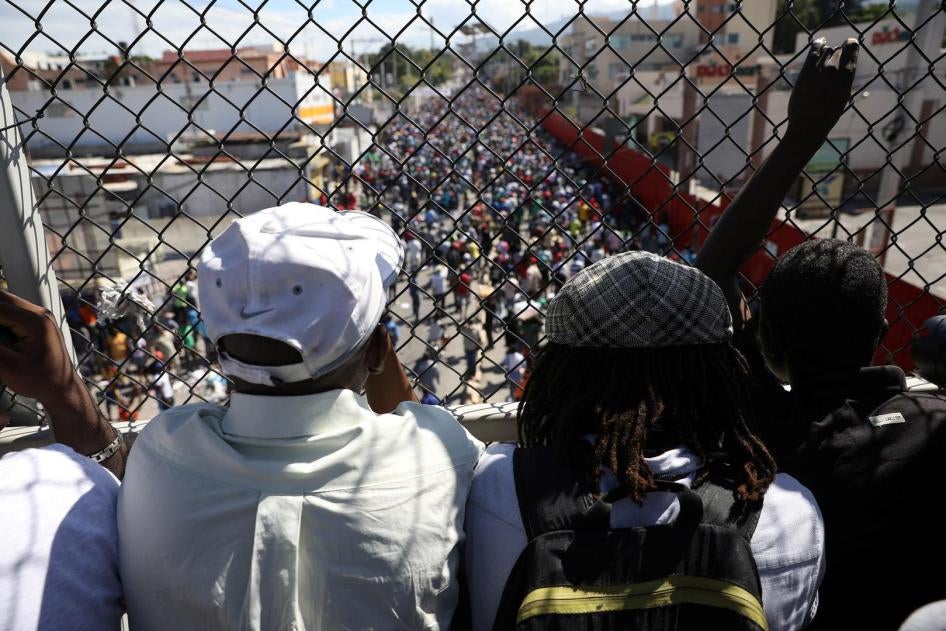 Men look at protesters marching to demand an investigation into what they say is the alleged misuse of Venezuela-sponsored PetroCaribe funds, in Port-au-Prince, Haiti, November 18, 2018. 