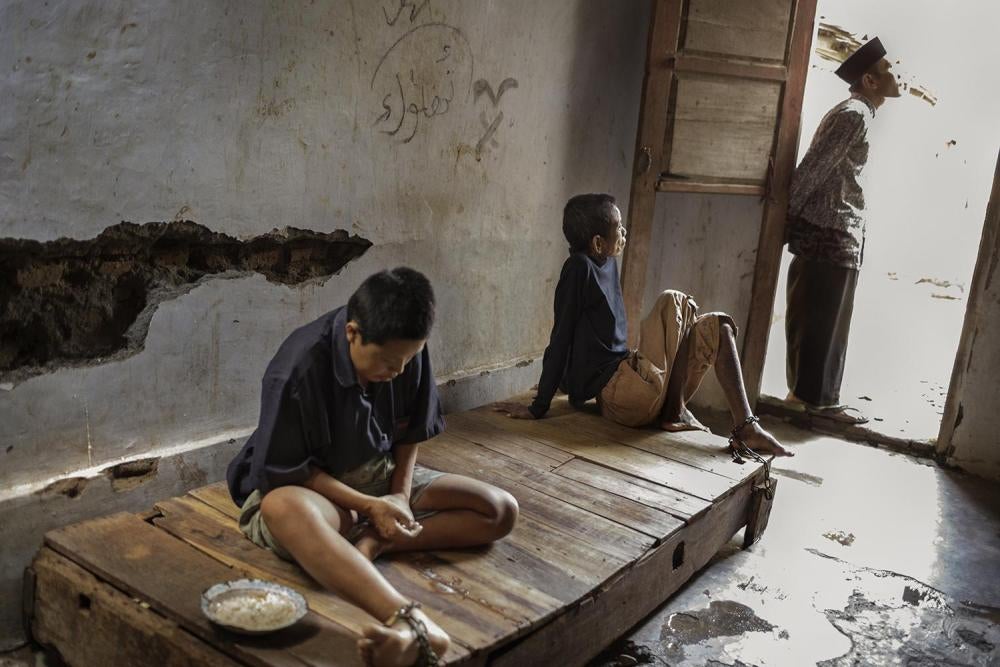 Two residents at the Bina Lestari healing center in Brebes, Central Java, are chained to a wooden platform bed while an Islamic faith healer stands nearby. At the center, all residents are chained and receive traditional “healing” through prayer, consumpt