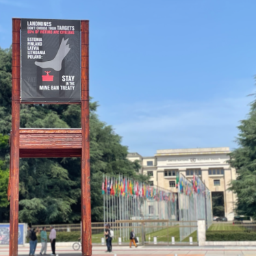 A banner hangs on the Broken Chair sculpture stating "Landmines don't choose their targets. 85% of victims are civilians. Estonia, Finland, Latvia, Lithuania, and Poland stay in the Mine Ban Treaty."