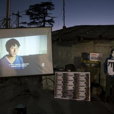 An exile Tibetan wearing a mask in the likeness of Tashi Wangchuk, stands next to a screen projecting a New York Times video during a street protest demanding his release, in Dharmsala, India. © 2018 AP Photo/Ashwini Bhatia