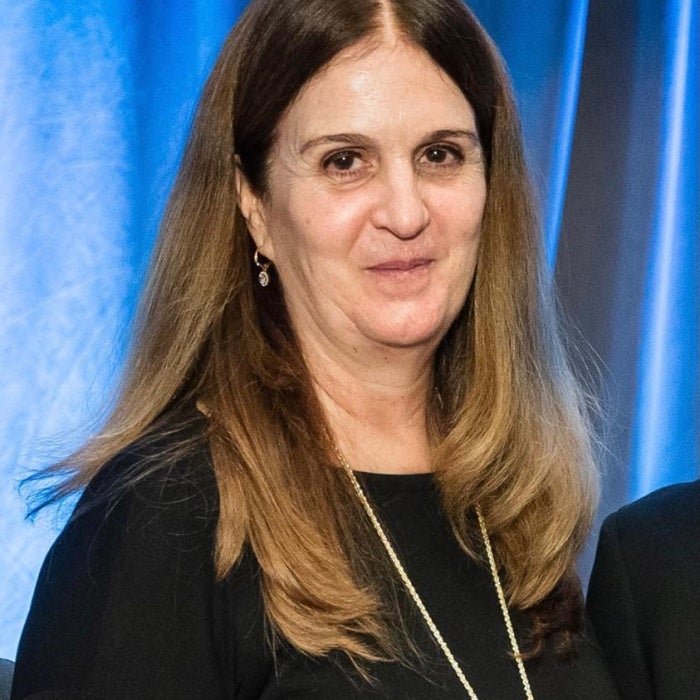 A woman with brown hair and pale skin wearing a black dress and long necklace smiles as she stands in front of a blue curtain